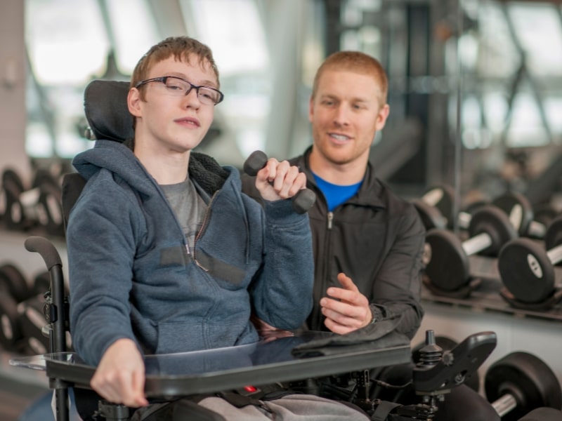 Young man in a wheelchair lifting weights with a trainer - disability help through physical training.