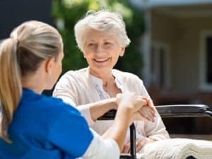 An elderly woman with short white hair sits in a wheelchair, smiling and looking at a younger woman with blonde hair in a ponytail wearing blue scrubs. The younger woman holds the elderly woman's hands, suggesting a supportive interaction related to navigating disability services. They appear to be outdoors, possibly in a garden or near a residential building, indicating a home care or assisted living setting.