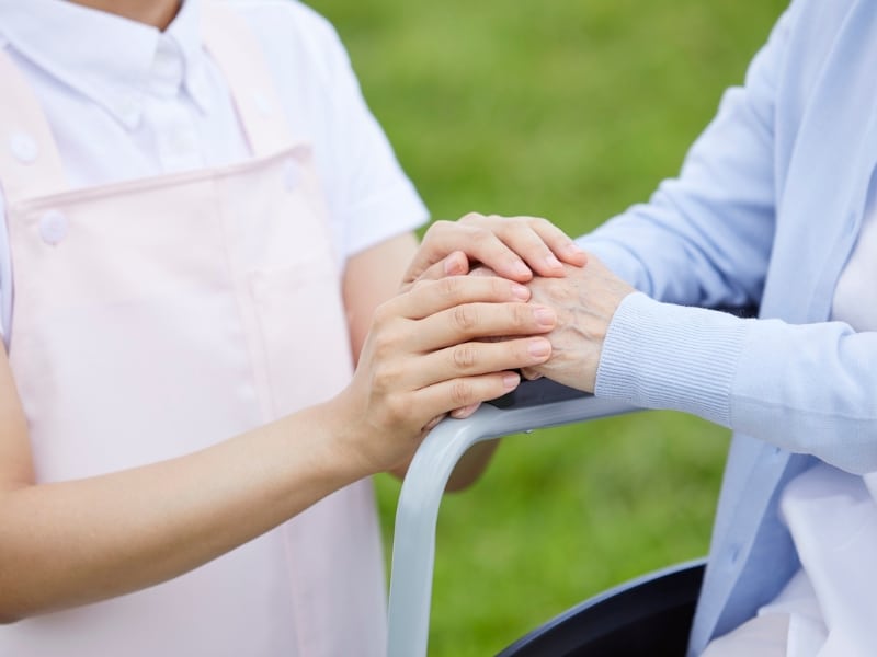 A close-up shows two pairs of hands clasped together on the handle of a mobility aid, possibly a walker or wheelchair, suggesting someone is receiving support to assist personal activities potentially under the NDIS.