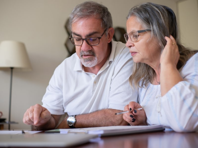 Older couple reviewing paperwork and discussing funding for disability services with serious concern at their kitchen table.