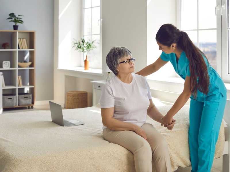 Support for special needs shown as a caregiver assists an elderly woman sitting on a bed in a home setting